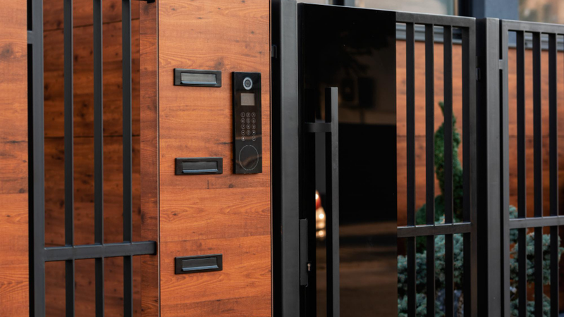 A modern home External Wooden Doors featuring a combination of warm wood paneling and sleek black metal fencing with a high-tech intercom system, representing external wooden doors for sale at Shield Doors and Windows.
